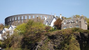 Un bâtiment historique en pierre, doté d’une tour ronde, entouré de maisons traditionnelles et d’une végétation luxuriante.