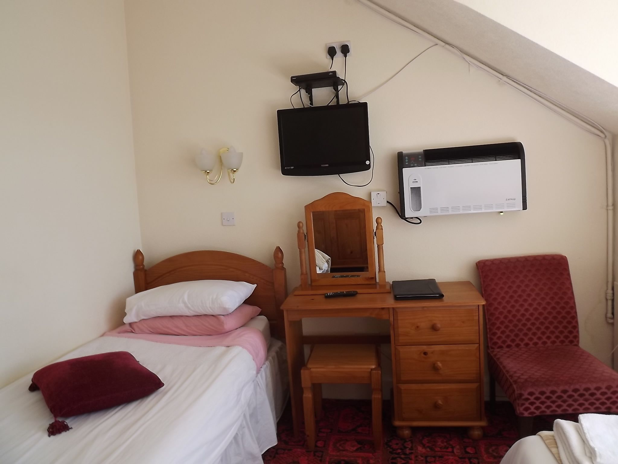 A bedroom with a bed, a wooden desk with a mirror, a chair, a TV, and an air conditioner.
