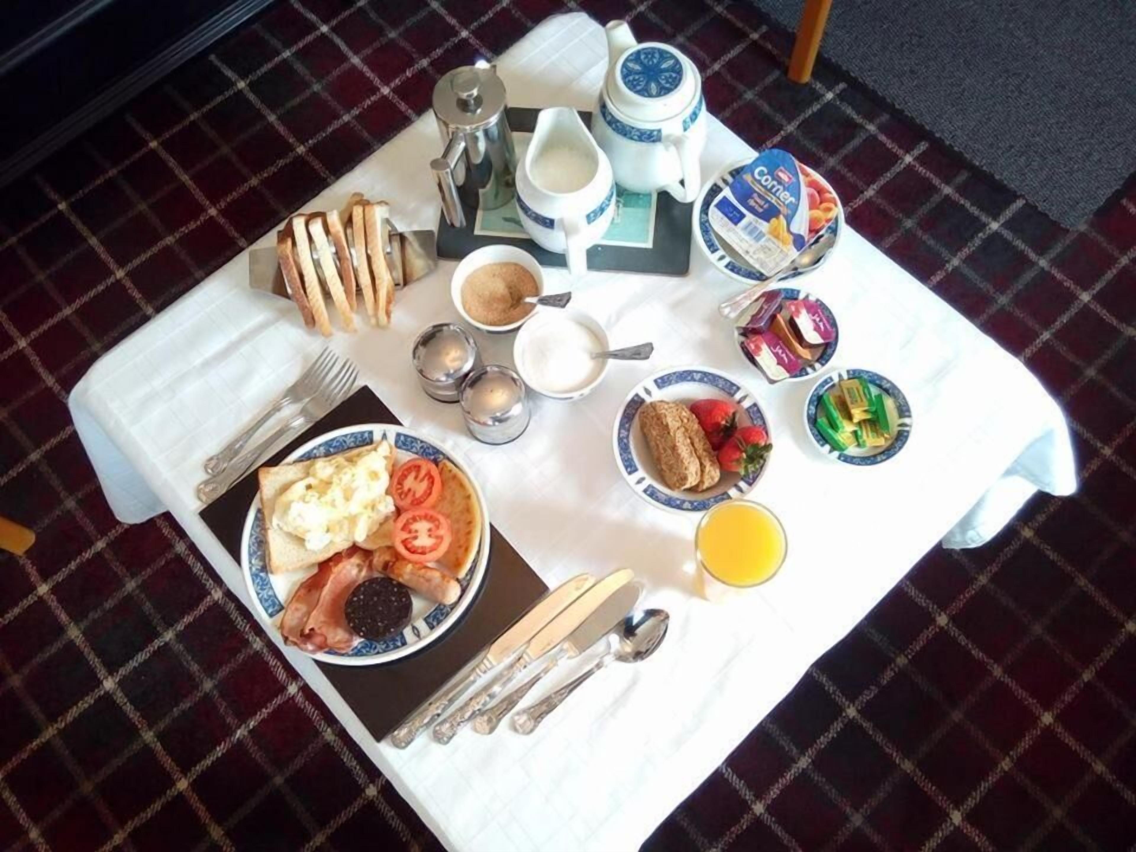 A breakfast table with a variety of food items, including toast, eggs, tomatoes, and a plate of pastries. There are also condiments, a coffee pot, and a jug of milk.