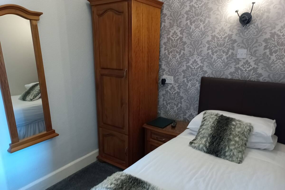 Quarto com cama, mesa de cabeceira, guarda-roupa, espelho e luminária na parede.
