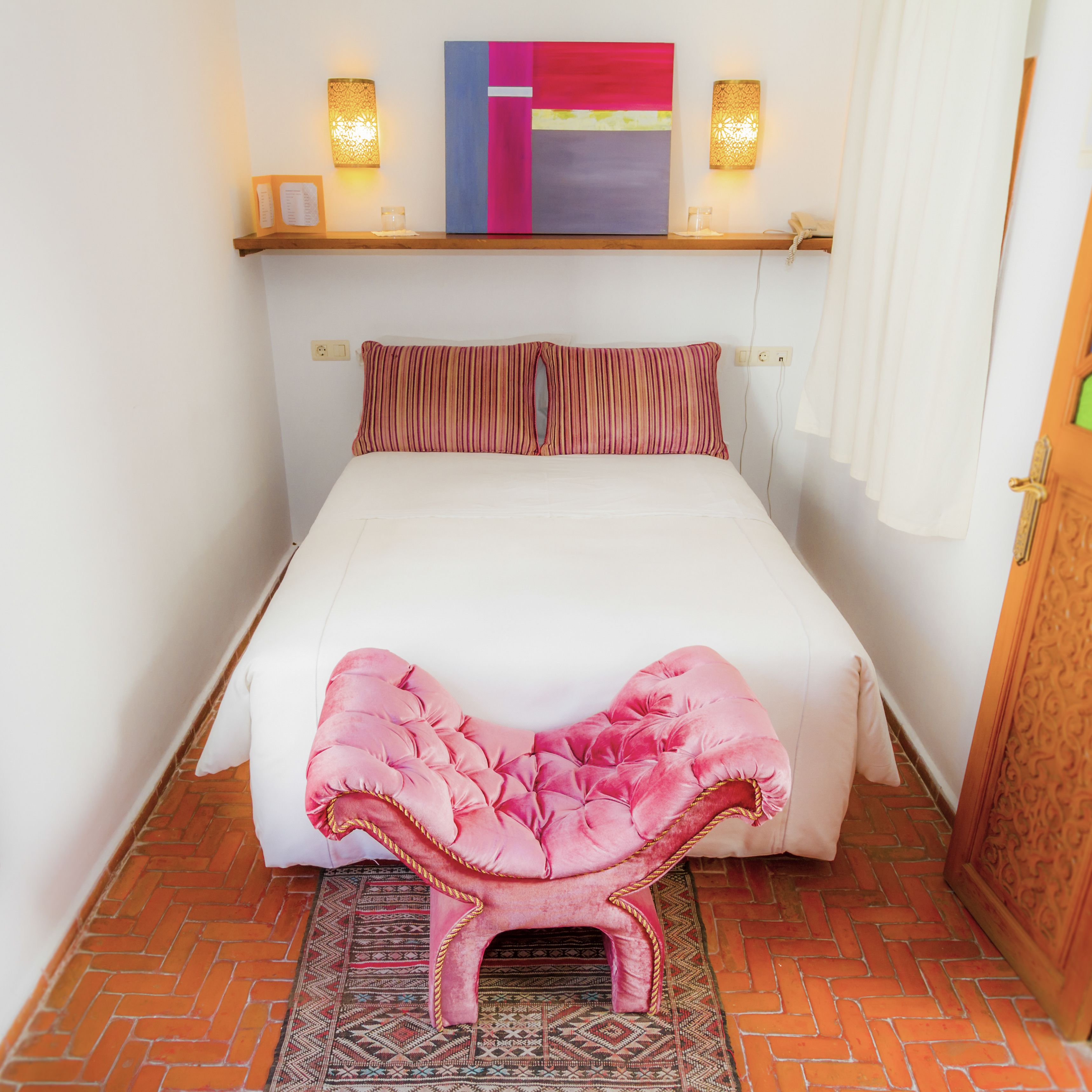 A bed with a white mattress and pink striped pillows, a pink ottoman, and a colorful abstract painting above the bed.