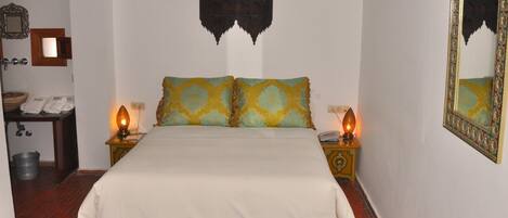Una cama bien hecha con una colcha blanca y dos almohadas con diseño estampado. Se observan dos mesitas de noche con lámparas, un adorno en la pared y un espejo con marco decorado.