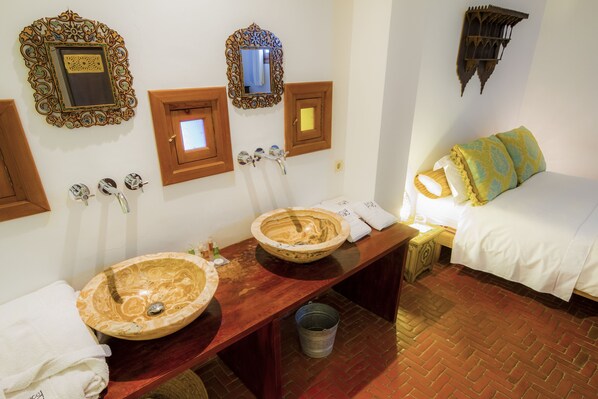 A bathroom with two large, ornate sinks, a wooden vanity, a bed with patterned pillows, and decorative wall mirrors.