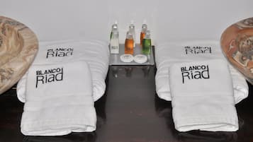 Duas toalhas brancas com o texto 'BLANCO Riad', um tabuleiro com quatro garrafas e um objeto com padrão marmorizado à esquerda.