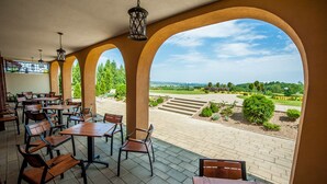 Ein Straßencafé mit Holztischen und -stühlen, Bögen und Blick auf einen angelegten Garten.