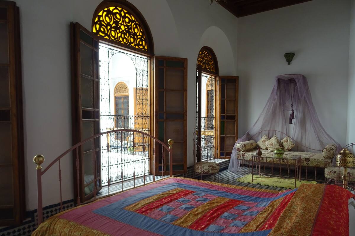 A bedroom with a large bed, a sofa, a canopy, and a view of the outside through ornate windows.
