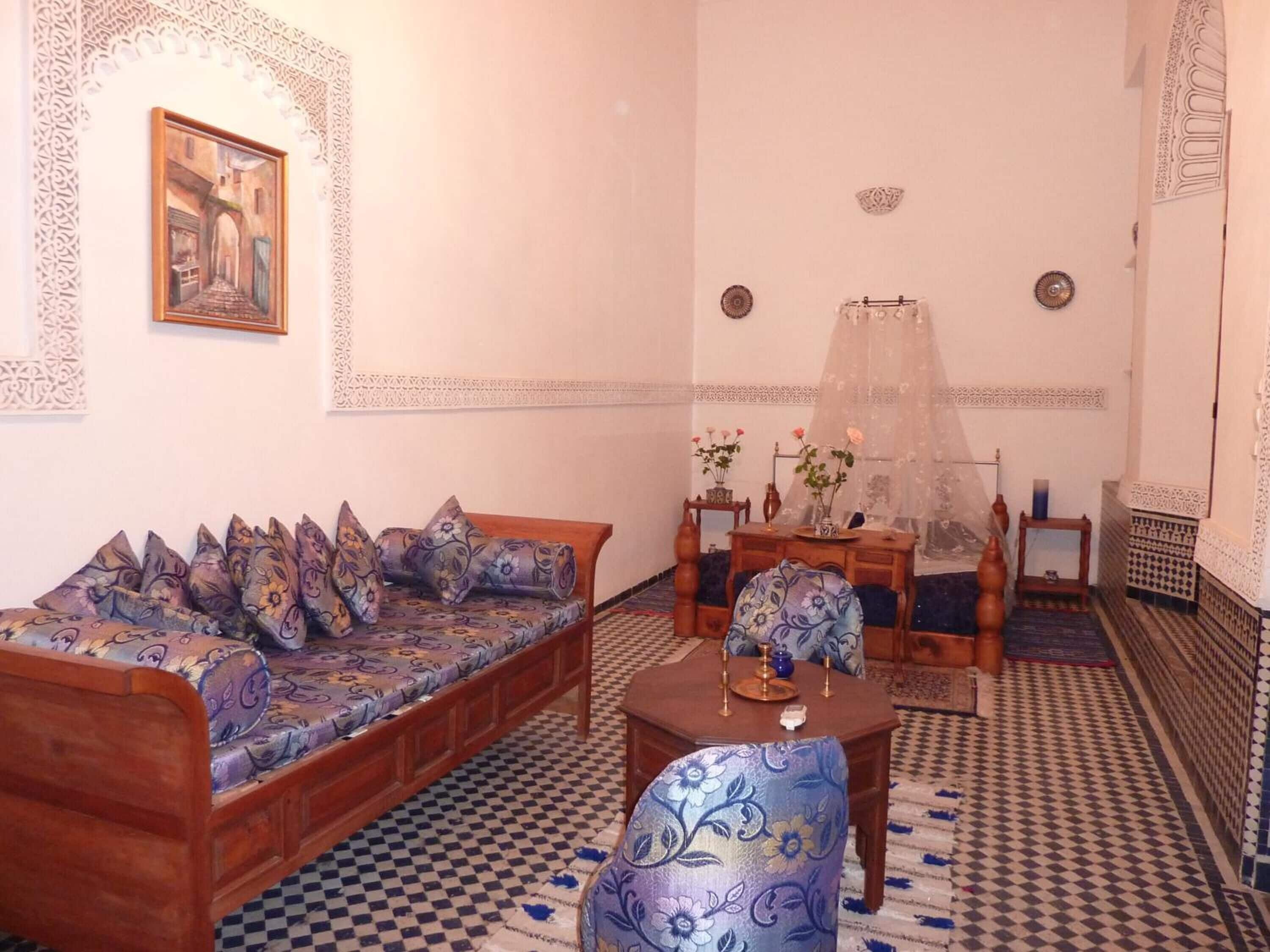 A traditional Moroccan room with a patterned checkered floor, a wooden sofa with floral cushions, a matching armchair, a small wooden table with a vase of flowers, and a decorative mirror on the wall.