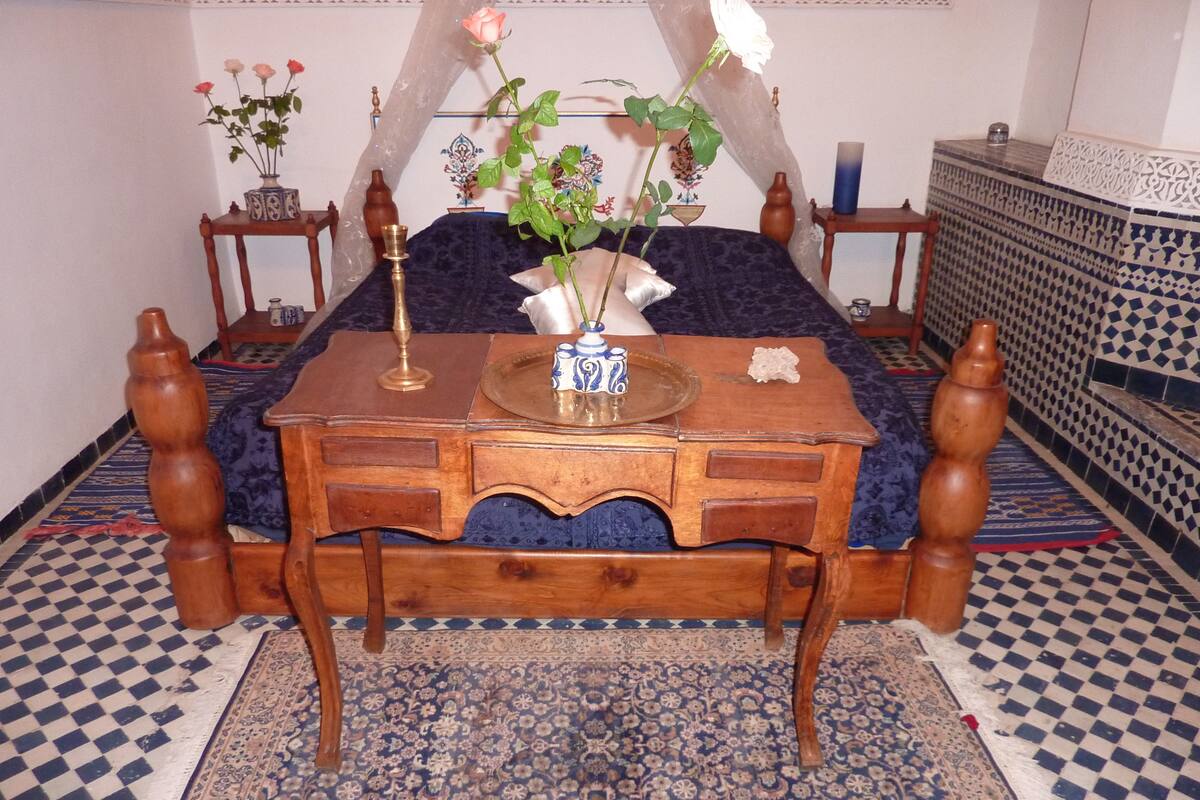 A traditional room with a wooden bed, a small wooden table with drawers, a decorative vase with flowers, and a patterned rug.