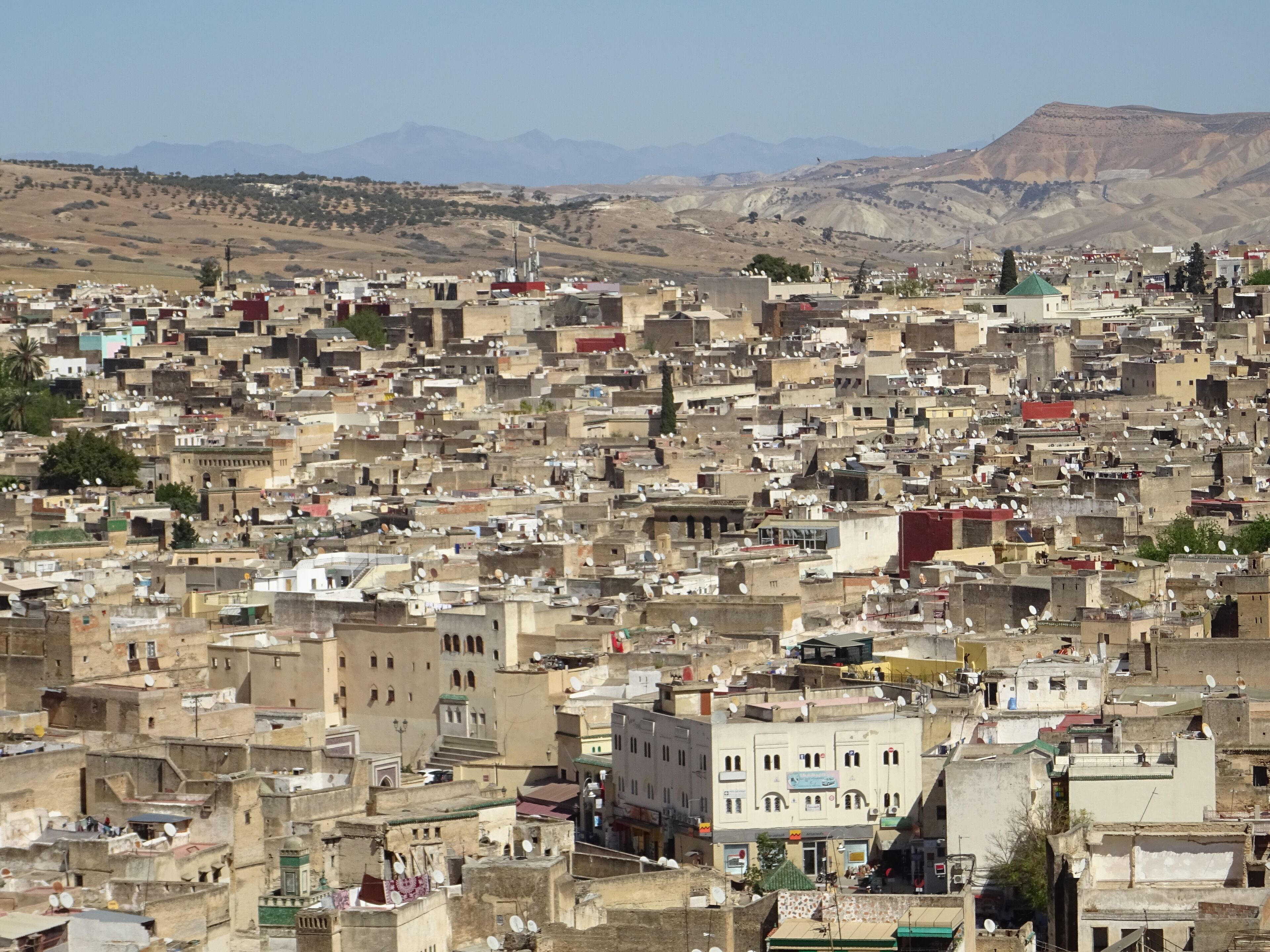 A densely packed urban area with numerous buildings, some with satellite dishes, and a mountainous backdrop.