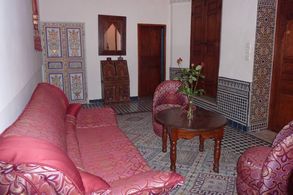 A traditional Moroccan interior with a patterned sofa, a small table with a vase, and a decorative cabinet.
