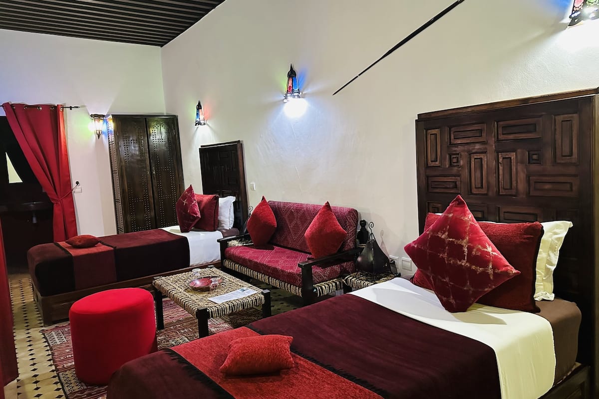 A room with a sofa, two single beds, a red ottoman, a small table with a bowl, and a wall-mounted light fixture.