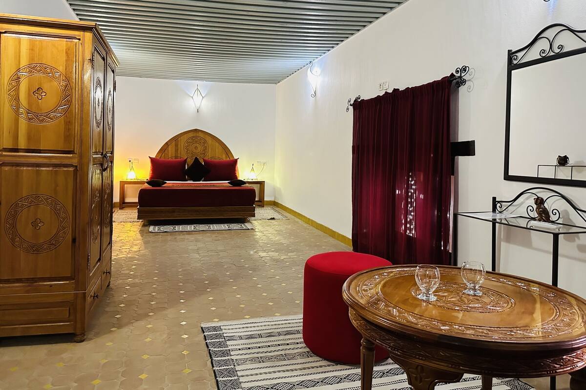 A room with a bed, a wooden wardrobe, a red ottoman, a round table with glassware, and a mirror on the wall.