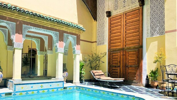A swimming pool with tiled edges, a lounge chair, and a large wooden door with intricate carvings. - Riad Zitouna (Fes)