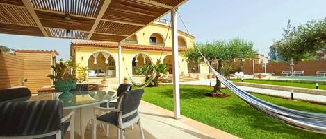 An outdoor patio with a glass table and chairs, a hammock, and a building with arched windows in the background.