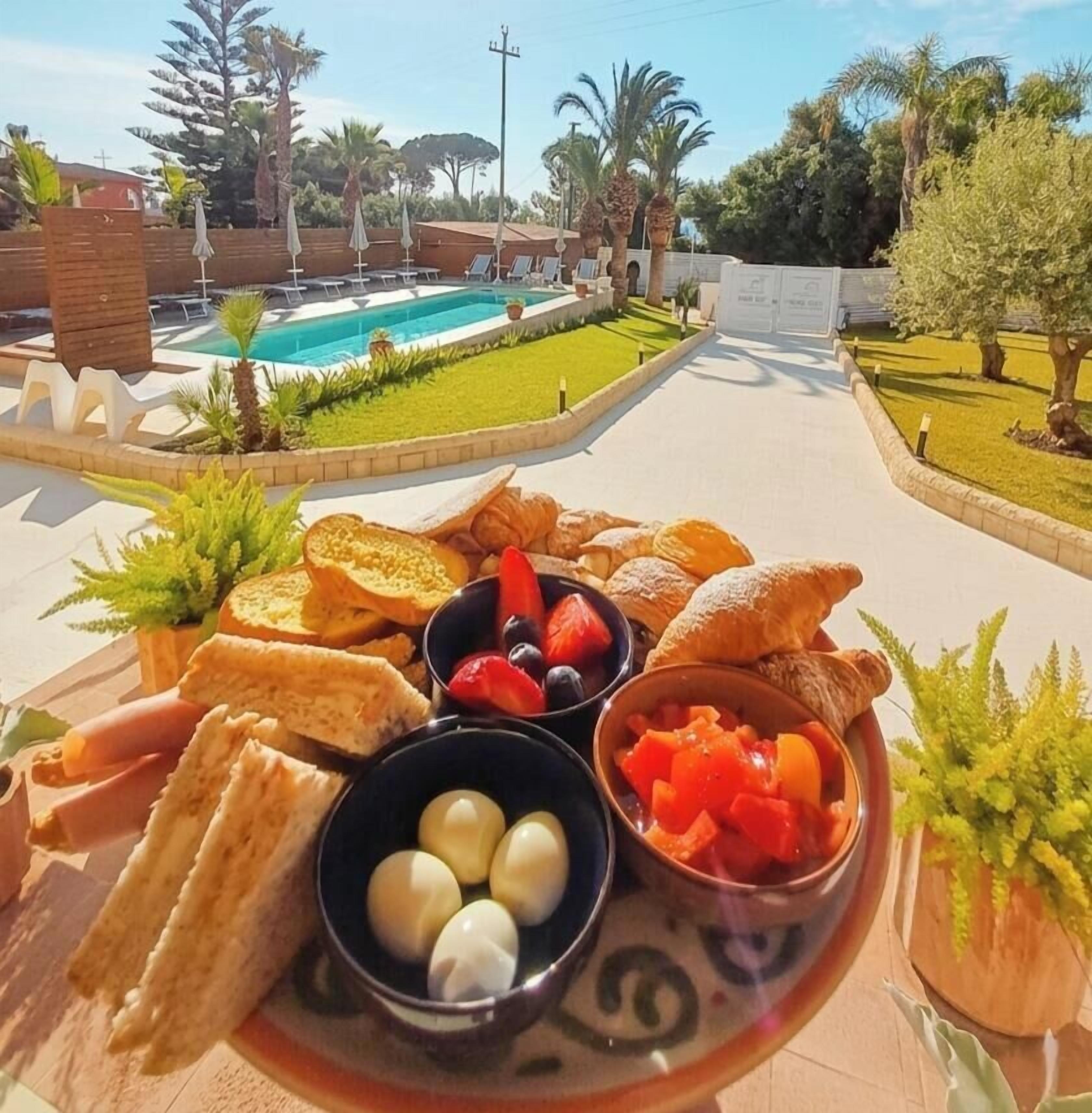 A breakfast spread with bread, fruits, and eggs on a tray, set outdoors with a pool and greenery in the background.