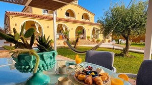 Una mesa de desayuno con croissants, frutas y jugo, ubicada al aire libre con un edificio al fondo.