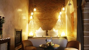A hotel room with a brick wall, a bed with white pillows, two armchairs, a small table with fruit, and wall-mounted lights. - Riad Kalaa (Rabat)