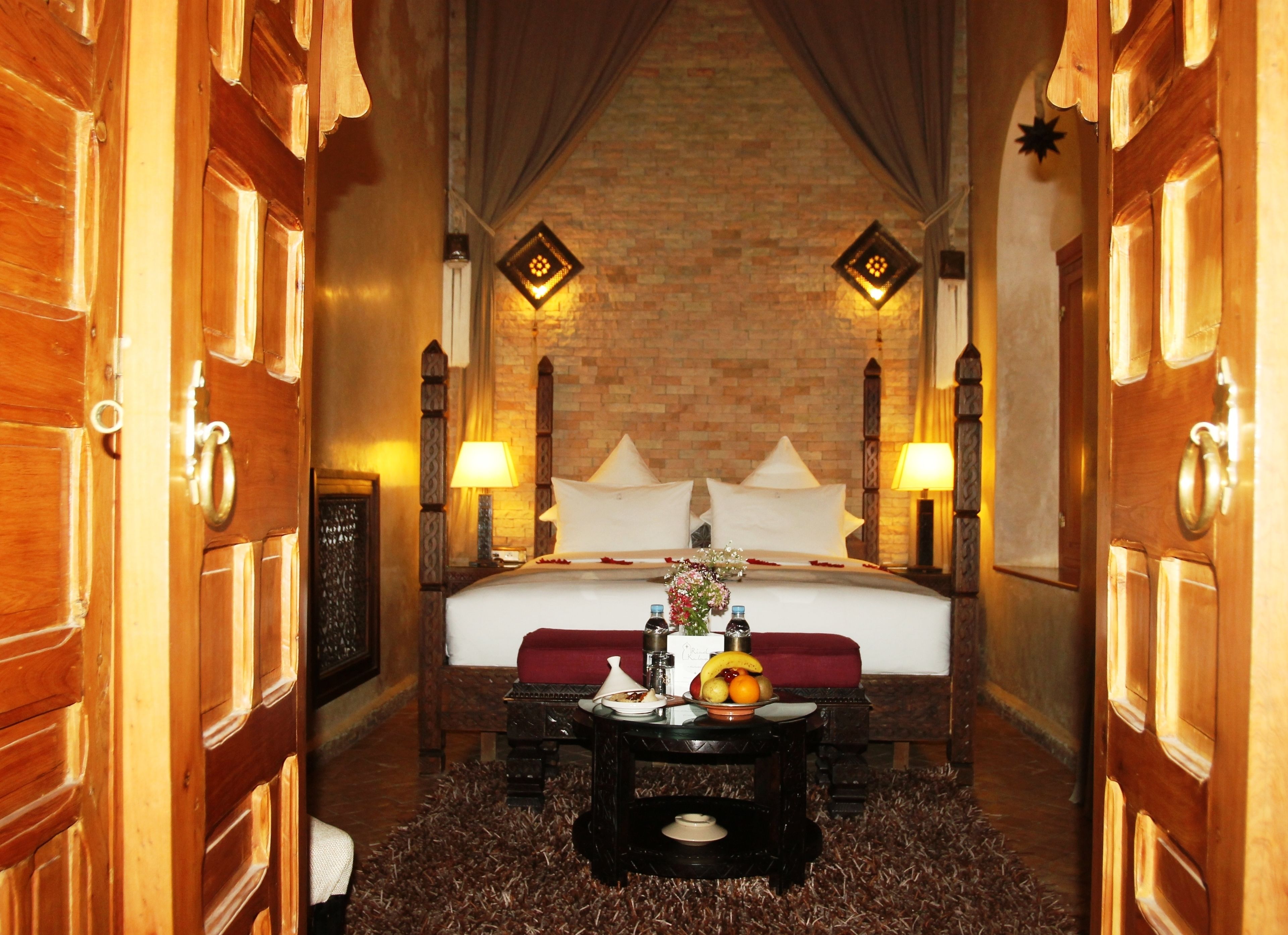 A hotel room with a large bed, wooden doors, a brick wall, and two wall-mounted lamps.