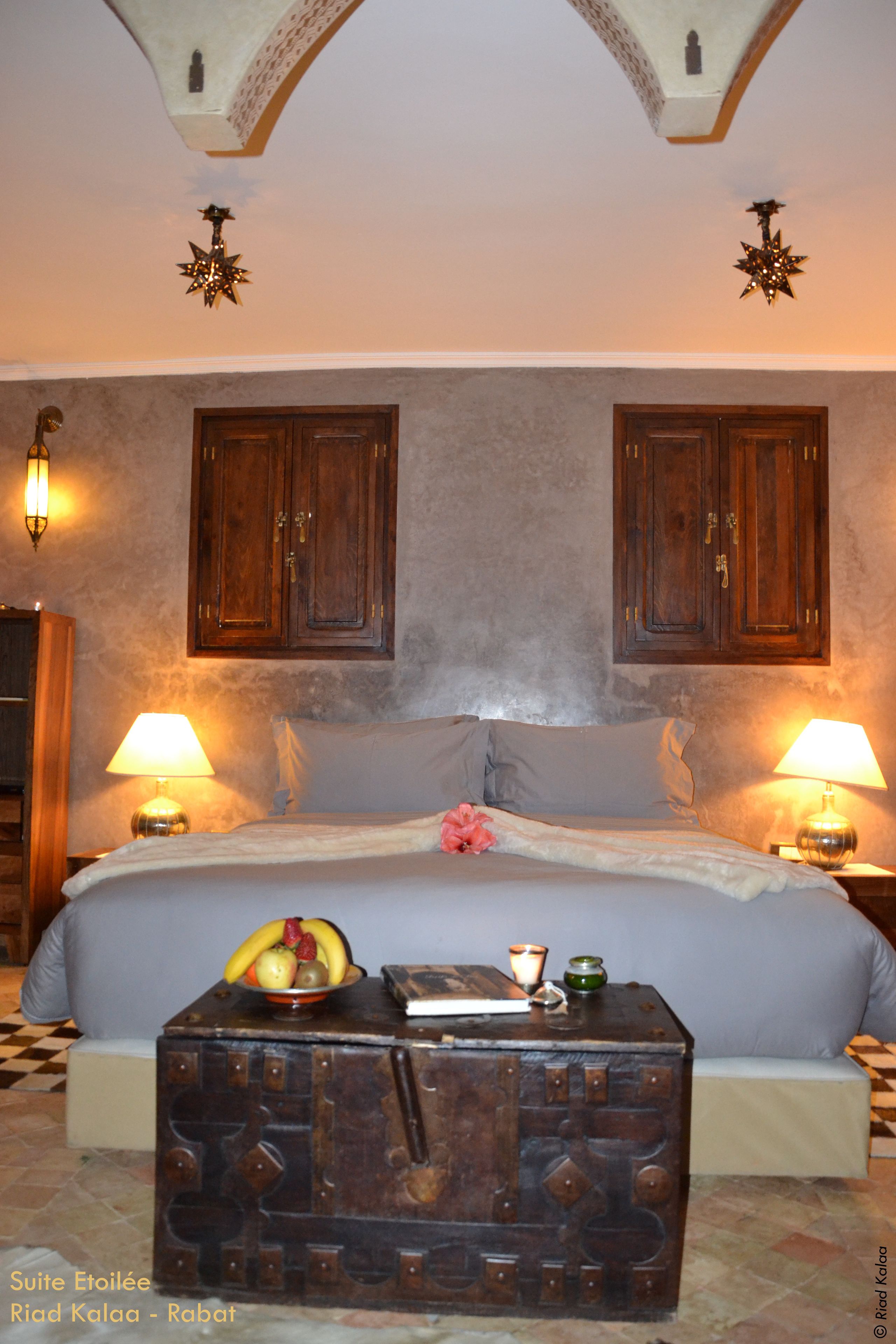 Une chambre à coucher avec un grand lit, deux armoires en bois, deux lampes fixées au mur, une commode décorative et une petite table sur laquelle se trouvent un panier de fruits et une bougie.
