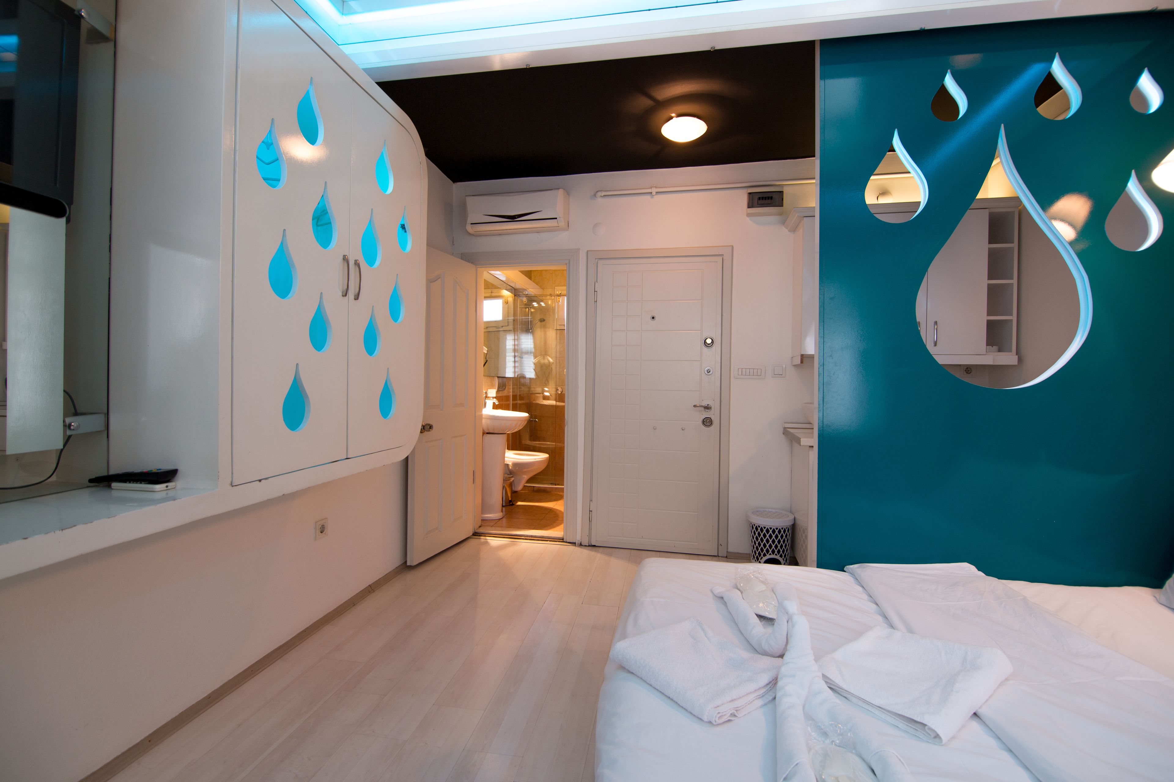 Quarto de hotel com cama, guarda-roupa decorado com gotas de água, banheiro com vaso sanitário e pia, e uma parede em tom de verde-água com padrão de gotas.