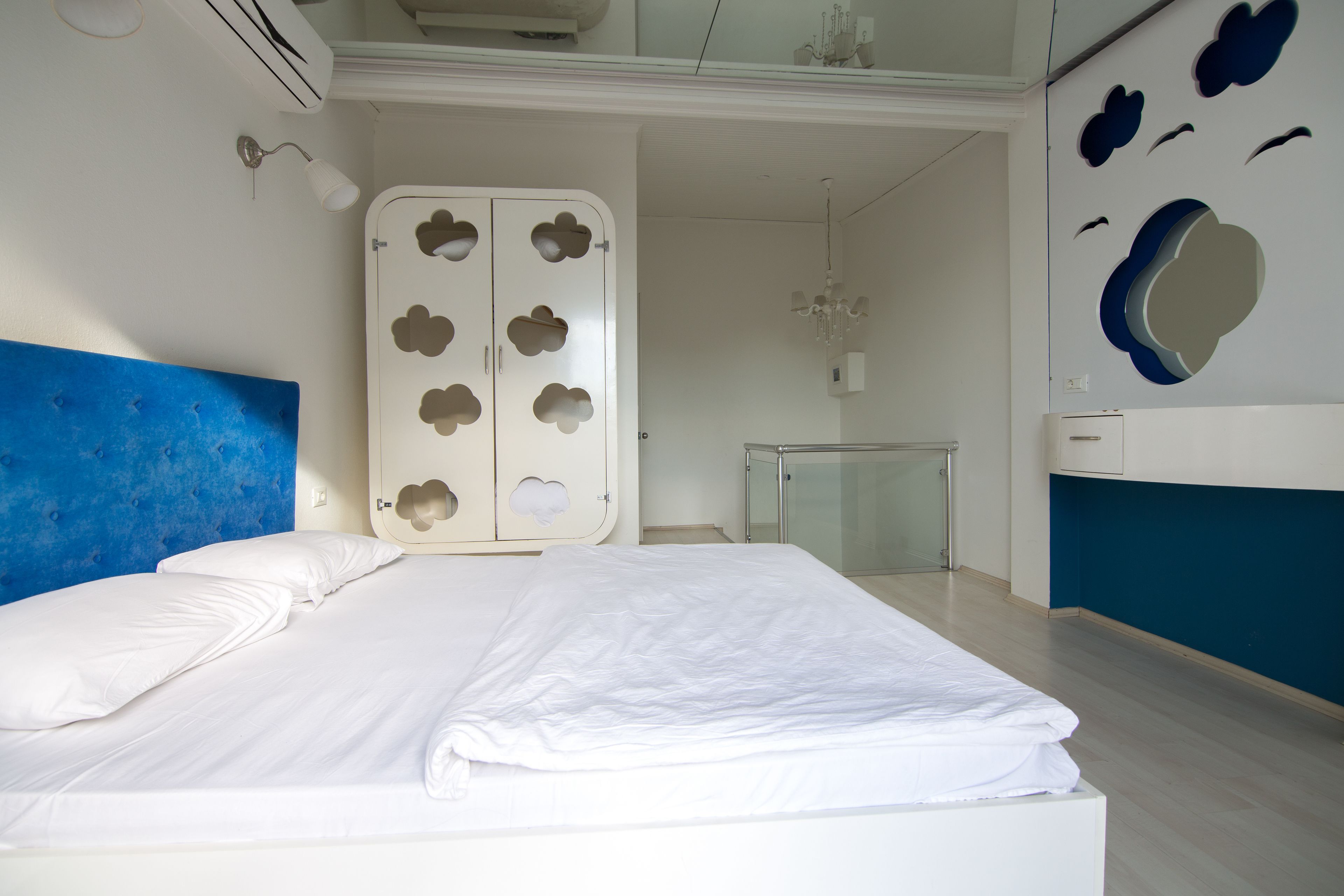 A bedroom with a bed, a wardrobe with cloud-shaped cutouts, a chandelier, and a bathroom with a glass door.