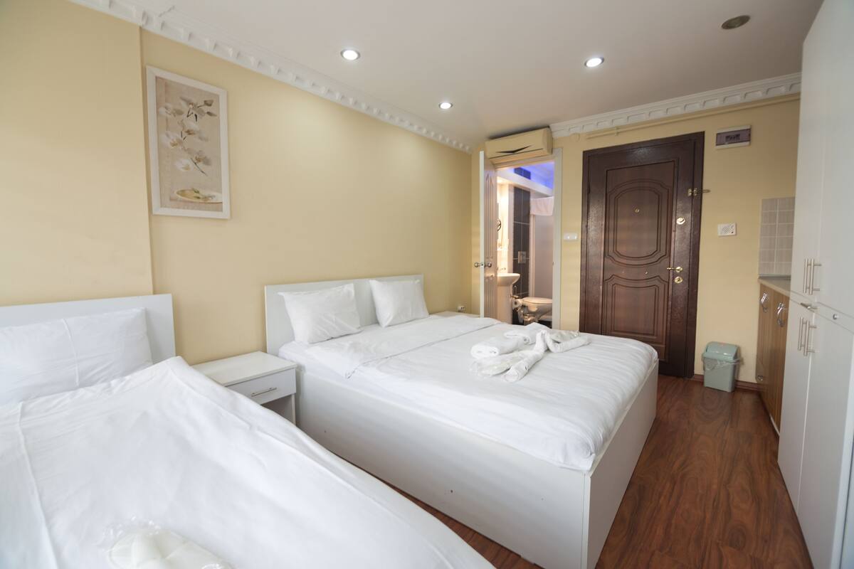 A hotel room with two beds, a bedside table, a small trash bin, and a door leading to a bathroom.