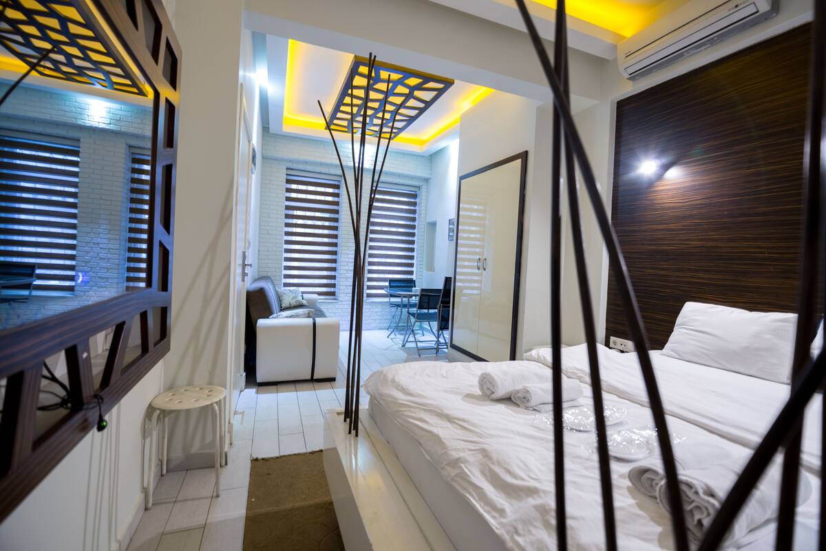 A modern hotel room with a large bed, a small seating area, and a view of the outdoor space.