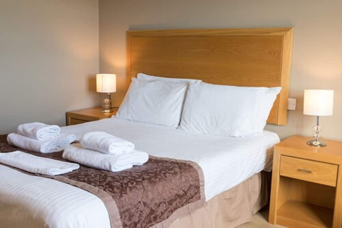Una cama bien hecha con sábanas blancas y una manta marrón, dos lámparas de noche y una mesita de noche de madera con un cajón.