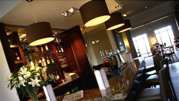 A bar area with a counter, bar stools, and a large vase of flowers. There are hanging lamps and a view of a dining area in the background. - Purdy Lodge (Belford)