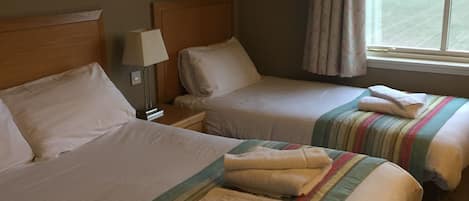 A hotel room with two beds, each with a headboard, a bedside table with a lamp, and a window with curtains.