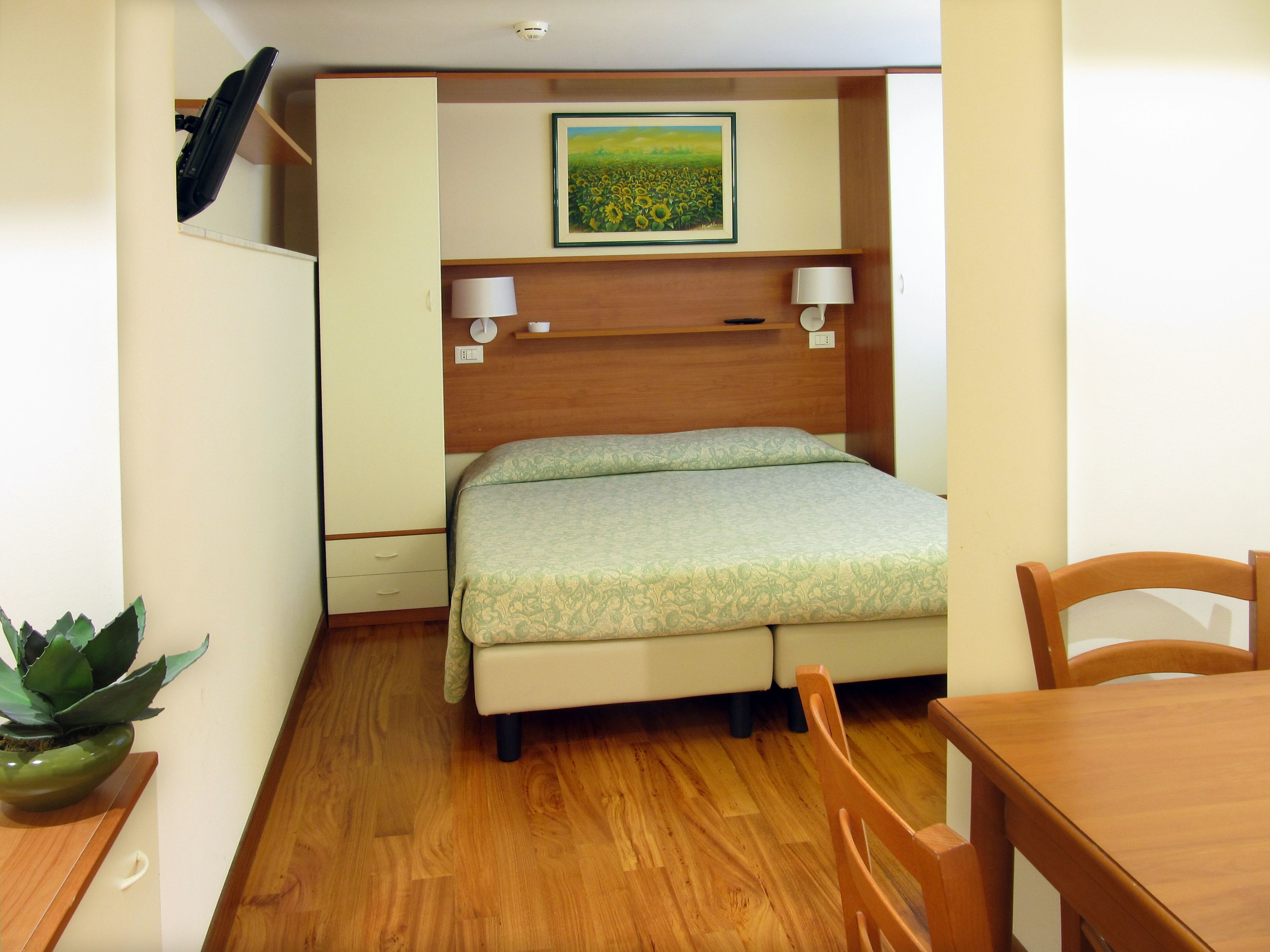 A compact room with a bed, a small table, a chair, a potted plant, and a wall-mounted shelf with a picture.