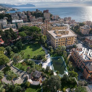 An aerial view of a coastal cityscape with a prominent building featuring a rooftop pool and lush surrounding greenery. - Rex Hotel Residence (Genoa)