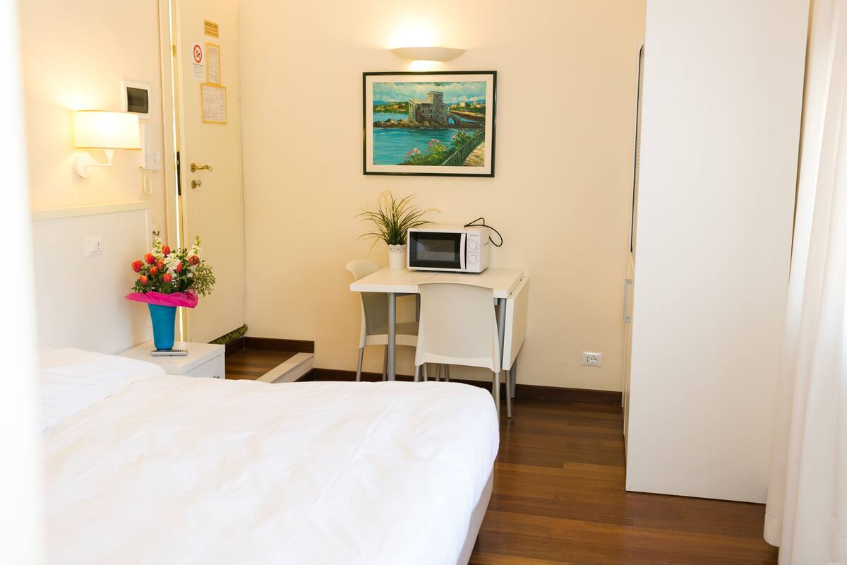 Une chambre d’hôtel avec un lit, un bureau équipé d’un ordinateur, une petite plante en pot et un tableau accroché au mur.