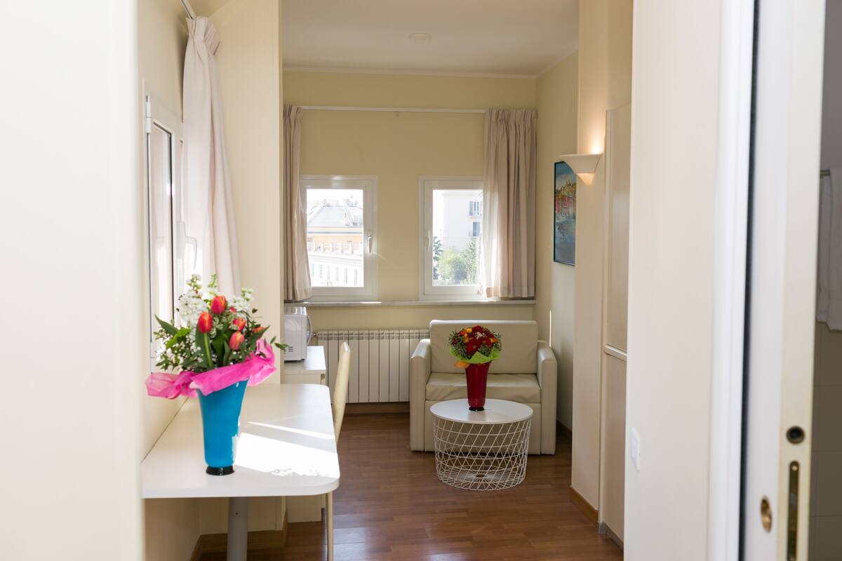 Un couloir étroit comprenant une table à manger, une chaise, une petite table ronde ornée d’un vase de fleurs et un canapé.