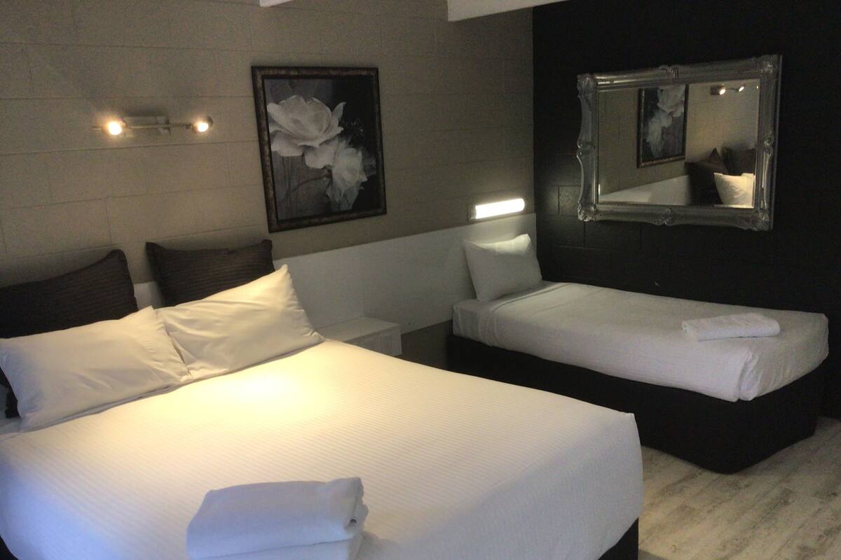 Hotellihuoneessa on suuri sänky, pienempi sänky, yöpöytä, peili ja seinällä oleva kehystetty kuva.