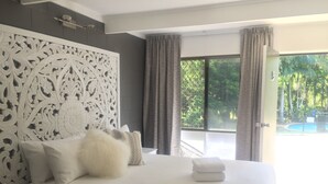 Ein Schlafzimmer mit einem großen Bett, weißer Bettwäsche, einem dekorativen Kopfteil und Blick auf einen Swimmingpool durch die geöffnete Tür.