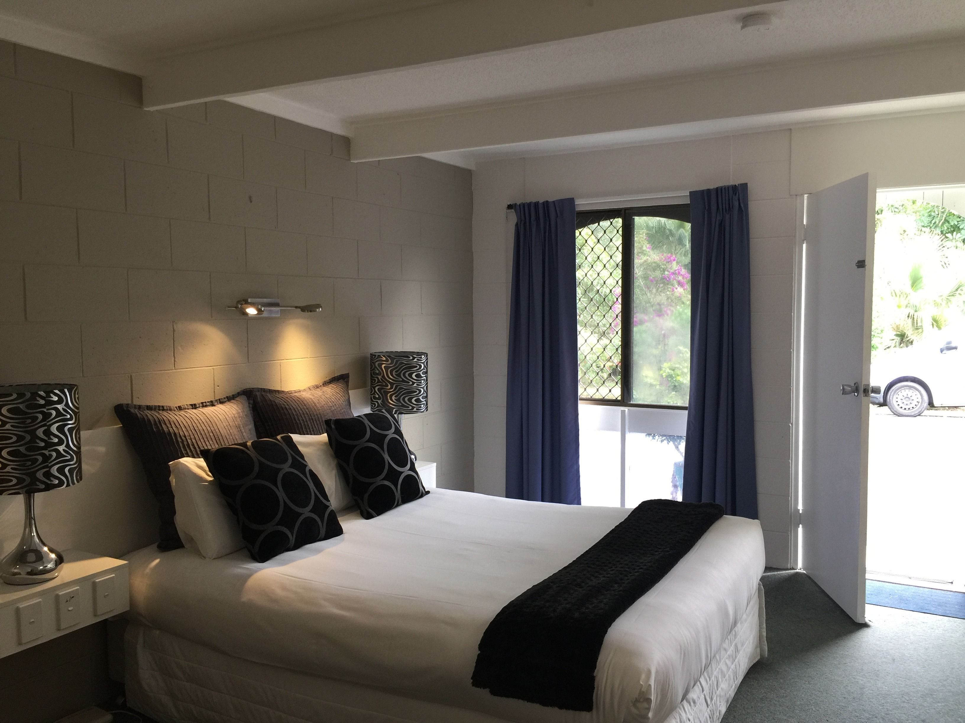 A bedroom with a bed, two bedside lamps, a window with curtains, and a door leading outside.