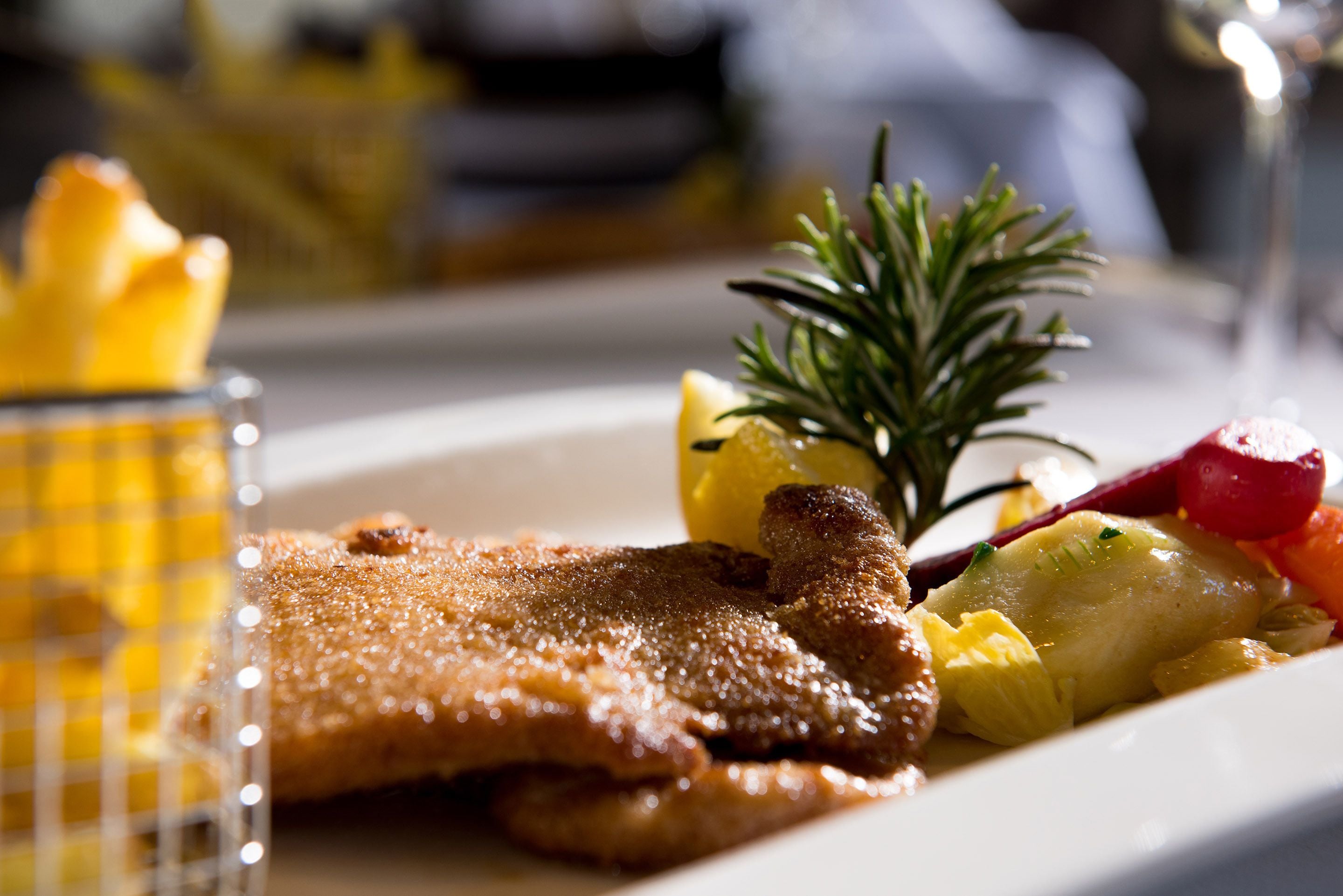 Un plato con un trozo de comida frita, posiblemente un schnitzel, acompañado de una ramita de romero y algunas verduras.
