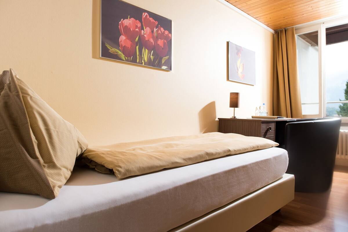 Quarto de hotel moderno com sofá-cama, escrivaninha e uma cadeira. Há duas pinturas na parede e uma janela com cortinas.