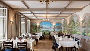 A restaurant with multiple tables and chairs, large windows, and a mural of a landscape on the wall. - Jungfrau Hotel (Wilderswil)