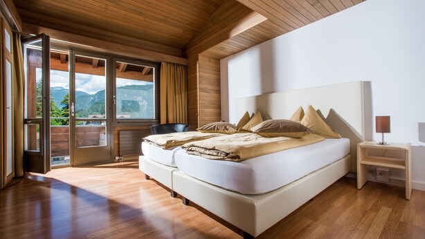 A bedroom with a large bed, wooden flooring, and a view of mountains through a sliding door. - Jungfrau Hotel (Wilderswil)