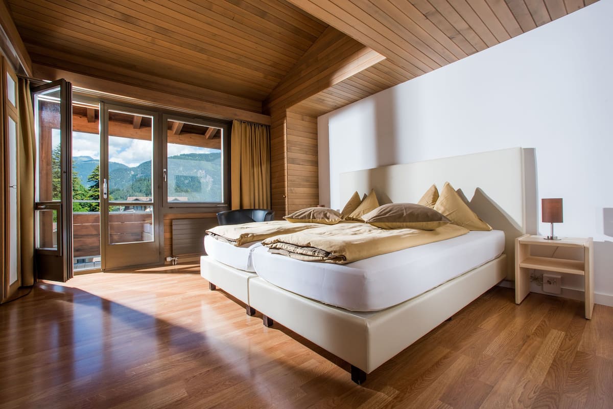Quarto com cama grande, piso de madeira e vista para montanhas através de uma porta de correr.