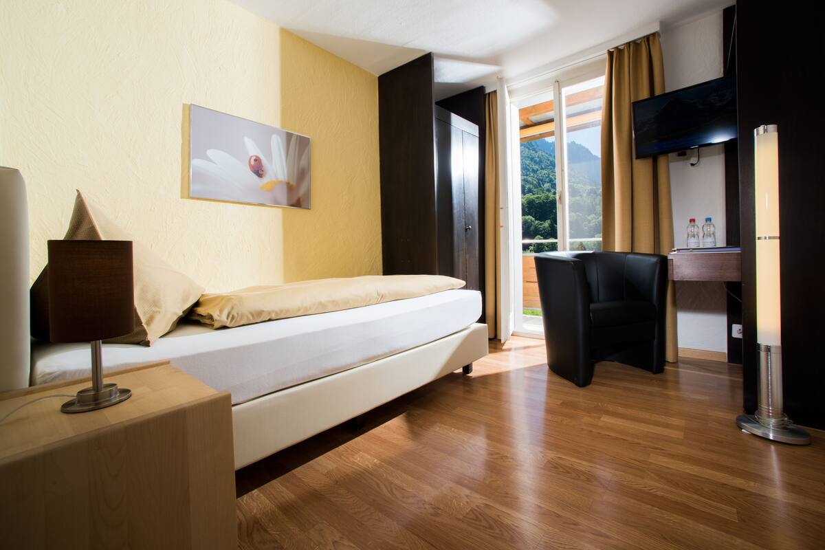 Quarto de hotel com uma cama, escrivaninha com luminária, uma cadeira e uma televisão.