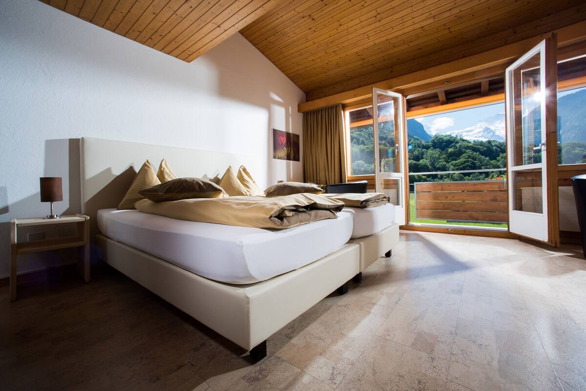 Quarto com cama grande, teto de madeira e vista para uma montanha através de janelas abertas.
