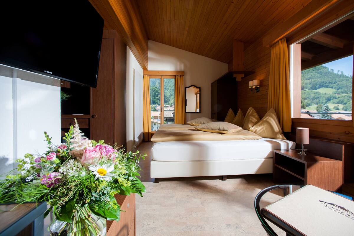 Quarto com cama, televisão, escrivaninha com luminária e janela com vista para o verde.