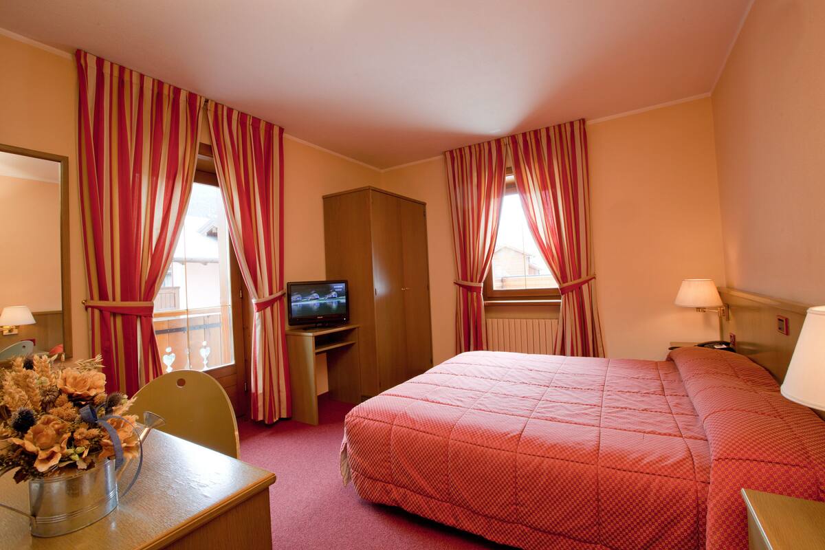 A hotel room with a bed, a desk with a television, a chair, and a vase of flowers.