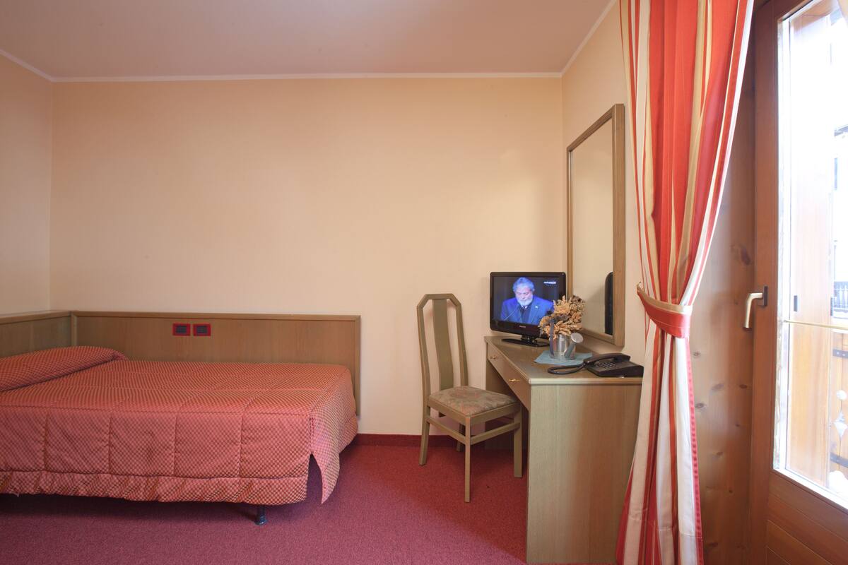A hotel room with a bed, a desk with a computer, a chair, and a window with curtains.