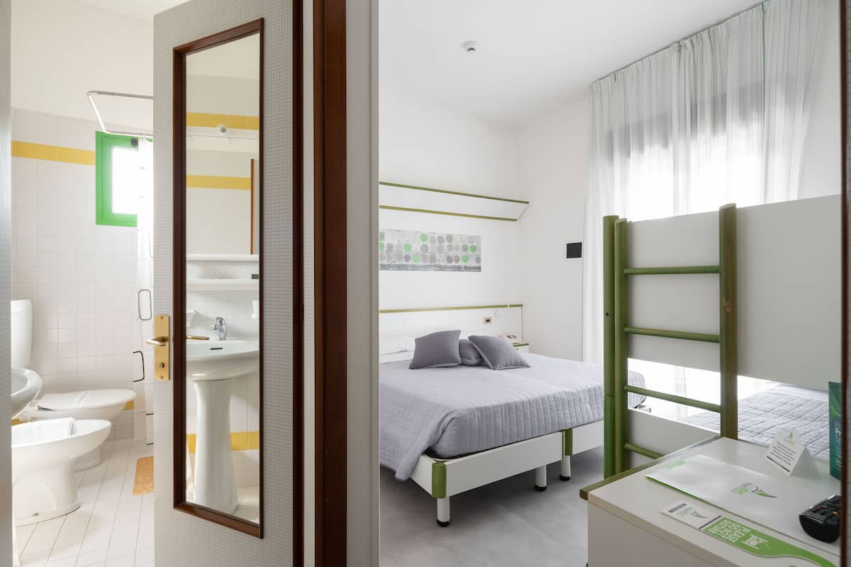 A modern hotel room with a bathroom, a bed with a green headboard, and a desk with a mirror.