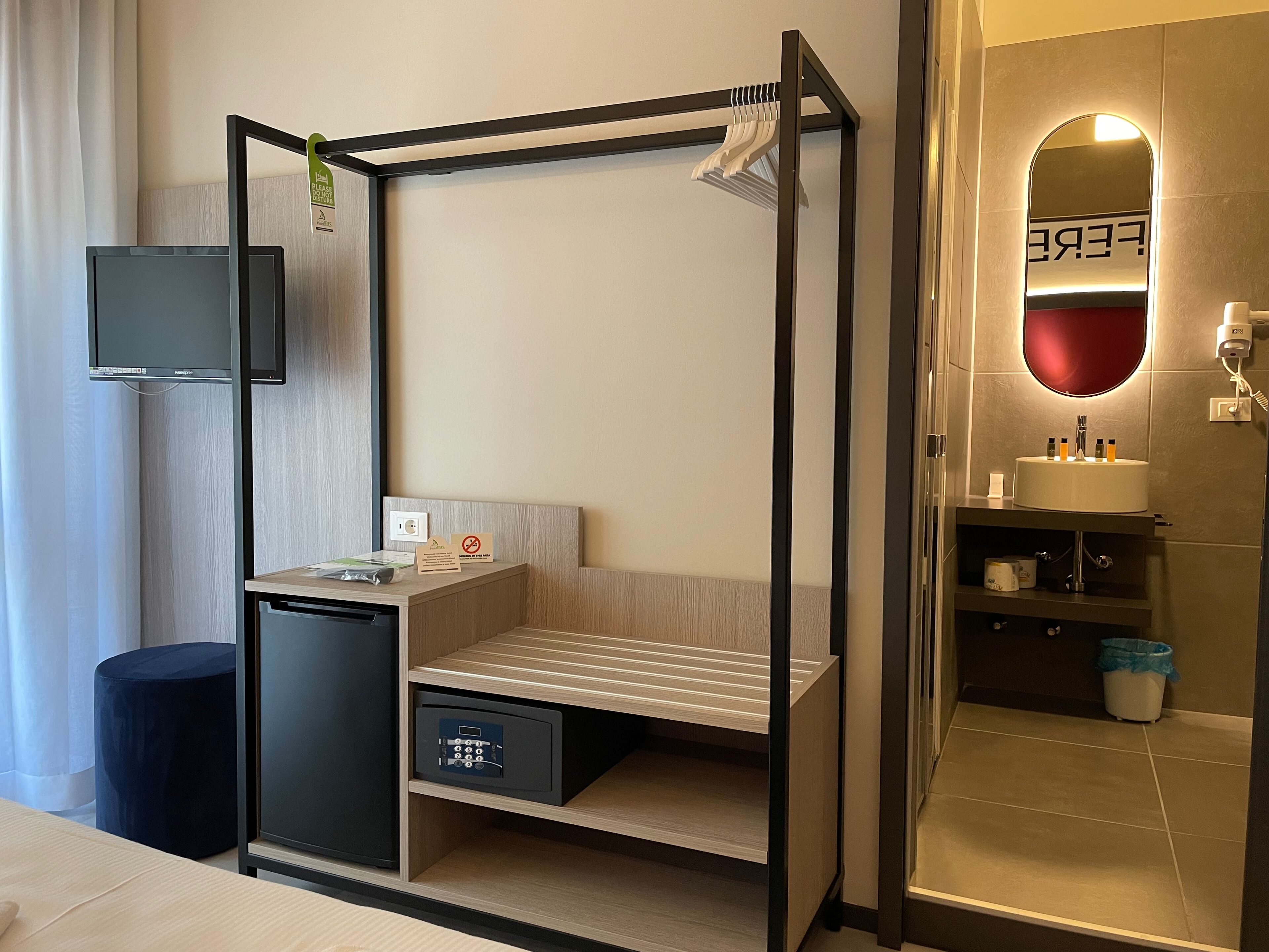Quarto de hotel pequeno com uma cama, uma escrivaninha, um banheiro compacto e uma televisão.