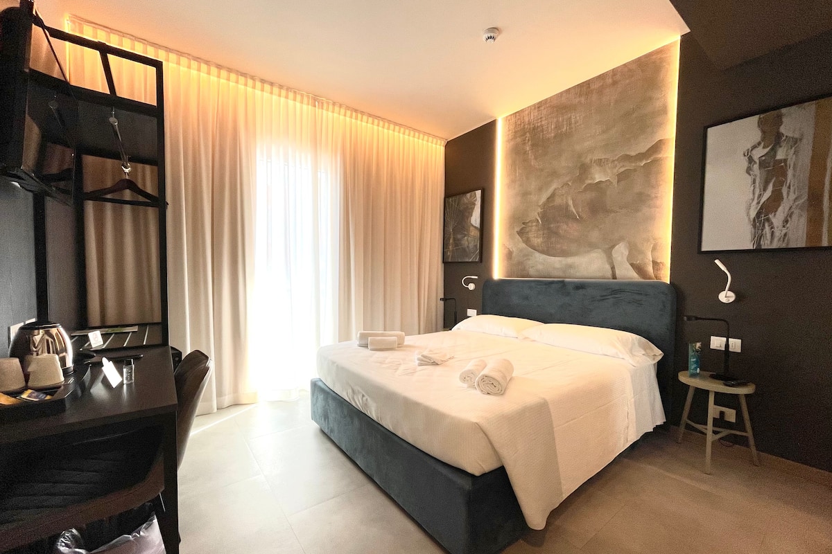 A modern hotel room with a large bed, a desk with a chair, and a small stool.